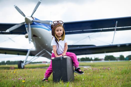 A little girl in a pilot's costume carries a retro suitcase and walks along the airfield. A child in a hat and glasses is going on a trip by plane