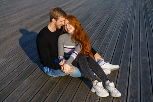 happy couple a guy and a girl with long red hair are sitting on a wooden deck in an embrace. Young man and woman of Caucasian ethnicity in casual clothes on a warm sunny day hugging