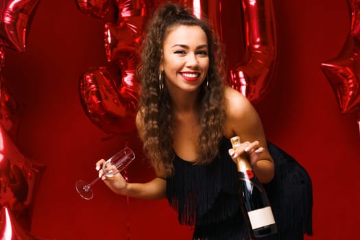 Beautiful woman posing on a red background with balloons holding a bottle of champagne in her hand. Cheerful girl of Caucasian appearance with wavy hair and bright makeup celebrates her 30th birthday