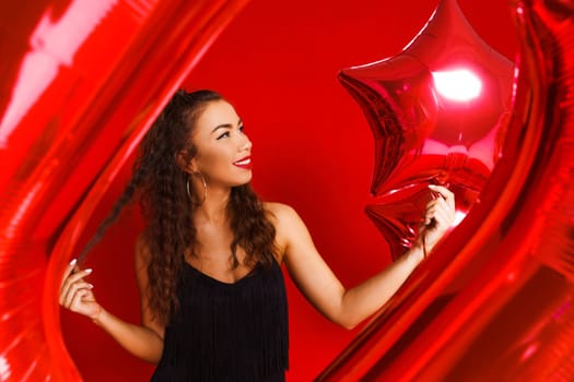 Portrait of a beautiful woman on a red background with red balloons posing in a black dress. The girl smiles with snow-white teeth on a holiday. Happy mood caucasian lady