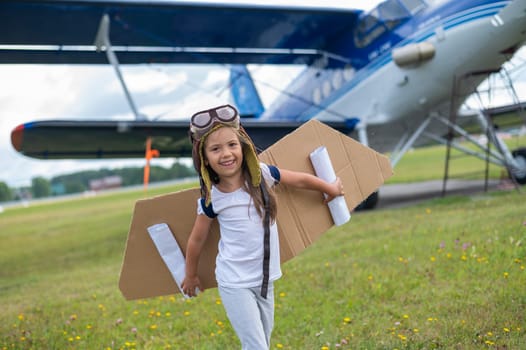 A little girl in a pilot's costume with cardboard wings runs on the lawn against the backdrop of the plane. A child in a hat and glasses dreams of flying on an airplane