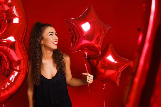 Portrait of a beautiful woman on a red background with red balloons posing in a black dress. The girl smiles with snow-white teeth on a holiday. Happy mood caucasian lady