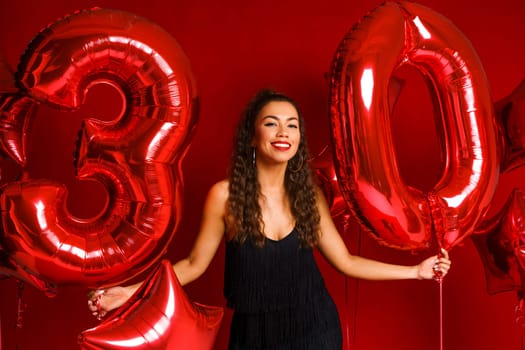 Portrait of happy 30 year old woman on red background with red balloons. A beautiful girl of Caucasian appearance is celebrating her anniversary. Balloons in the form of numbers 30 and stars.