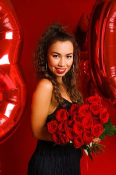 Beautiful young woman on a red background with red balloons and a bouquet of red roses. Cheerful and happy girl with bright red lipstick celebrates 30 years anniversary