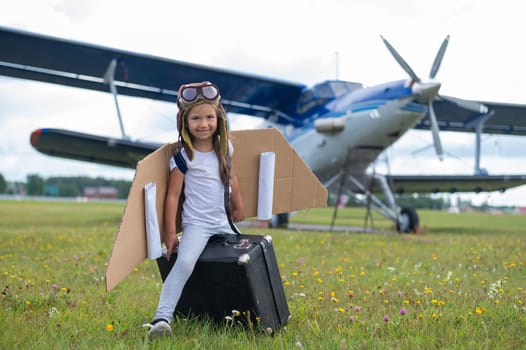 A little girl in a pilot's costume sits on a retro suitcase at the airport waiting for the departure of the flight. A child in a hat and glasses is going on a trip by plane