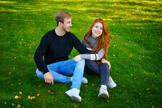 Cheerful young couple guy and red-haired girl of Caucasian ethnicity , are having fun together in the park on green grass on a sunny summer day in casual clothes