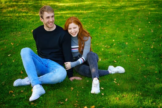 Cheerful young couple guy and red-haired girl of Caucasian ethnicity , are having fun together in the park on green grass on a sunny summer day in casual clothes