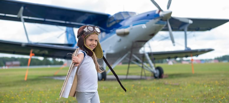 A little girl plays a pilot on the background of a small plane with a propeller. A child in a suit with cardboard wings dreams of flying.