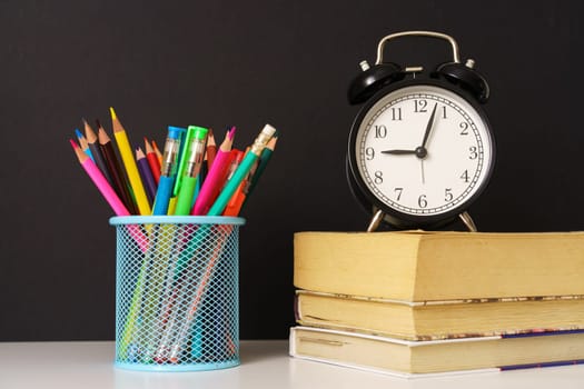 the alarm clock stands on the books and glass with pencils on black background, concept back to school