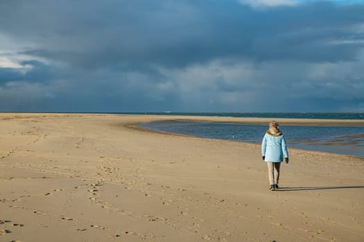 an adult woman in a blue coat walks on the wide sandy beach of Texel during a cloudy day with showers in the background