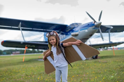 A little girl plays a pilot on the background of a small plane with a propeller. A child in a suit with cardboard wings dreams of flying.