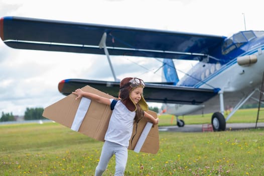 A little girl in a pilot's costume with cardboard wings runs on the lawn against the backdrop of the plane. A child in a hat and glasses dreams of flying on an airplane