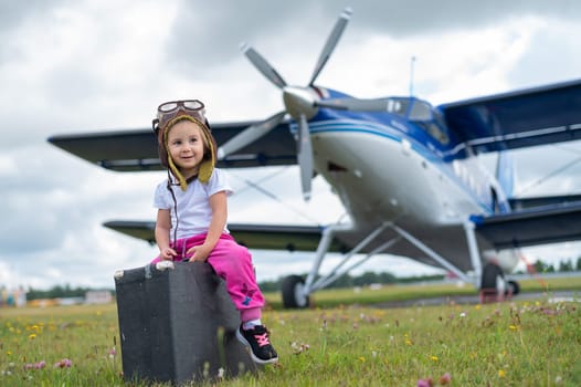 A little girl in a pilot's costume carries a retro suitcase and walks along the airfield. A child in a hat and glasses is going on a trip by plane