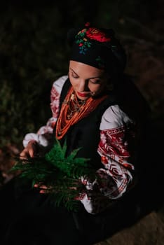 Witch woman collects herbs, ferns at night in Carpathian mountains forest. She in in traditional ukrainian handkerchief, national dress - vyshyvanka, ancient coral beads. Folk medicine concept.