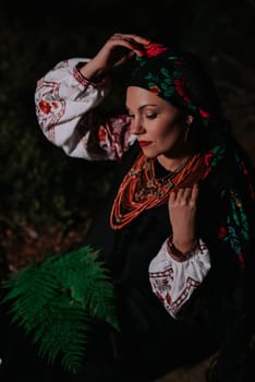 Portrait of ukrainian woman with fern at night in Carpathian mountains forest. She in in traditional ukrainian handkerchief, national dress - vyshyvanka, ancient coral beads.