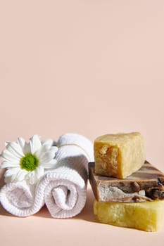 Spa set with two rolled white towels, chamomile flower and stack of organic natural soap bars with natural ingredients on isolated beige background. Vertical photo. Still life. Copy advertising space