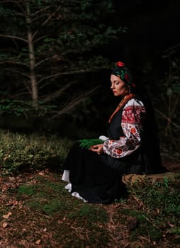 Witch woman collects herbs, ferns at night in Carpathian mountains forest. She in in traditional ukrainian handkerchief, national dress - vyshyvanka, ancient coral beads. Folk medicine concept.