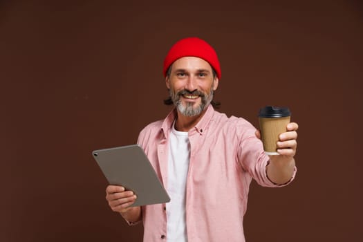 Work and coffee concept. Smiling mature man in casual clothing holds tablet in hand while offering craft cup with coffee in other hand. Happy mature freelancer caucasian man in red hat isolated on brown background. High quality photo