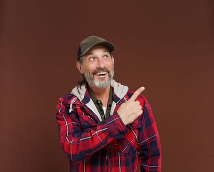 Cheerful American farmer or countryman with beard smiles and points his finger to copy space for advertisement or ad, looking with delight. Happy track driver concept. High quality photo