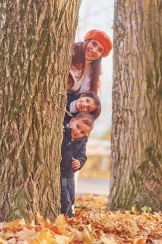 Hiding behind tree. Mother with her little son and daughter is having fun in the autumn park.