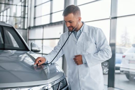 Medic in white coat is using stethoscope on the automobile.