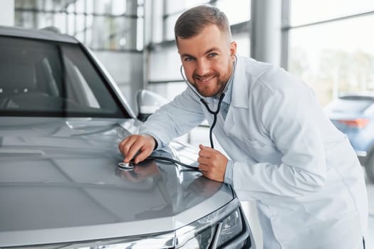 Medic in white coat is using stethoscope on the automobile.