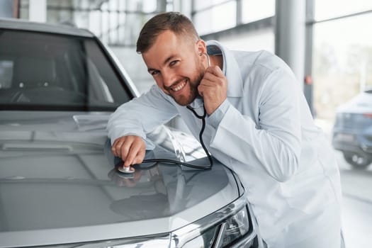 Medic in white coat is using stethoscope on the automobile.