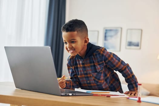 With laptop. Cheerful young boy is at home alone.