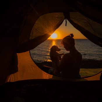 A woman and a dog in a tourist tent at sunrise. Camping with a pet.