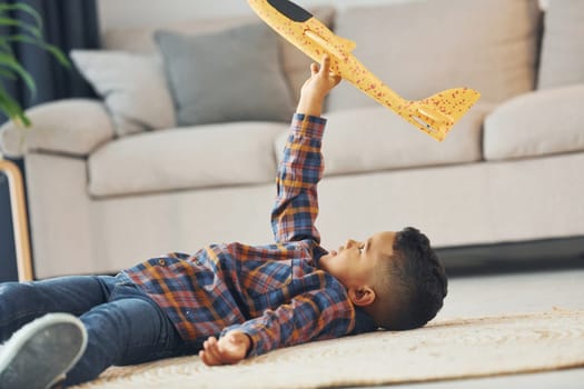Playing with yellow toy plane. Cheerful young boy is at home alone.