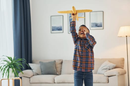 Playing with yellow toy plane. Cheerful young boy is at home alone.