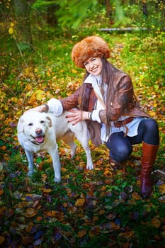 Girl in a leather jacket, a big red fox fur hat in the forest in autumn with white dog. A female model poses as fabulous royal huntress on nature hunt at photo shoot with animal pet