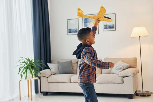 Playing with yellow toy plane. Cheerful young boy is at home alone.