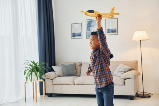 Playing with yellow toy plane. Cheerful young boy is at home alone.