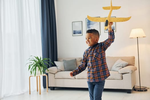 Playing with yellow toy plane. Cheerful young boy is at home alone.