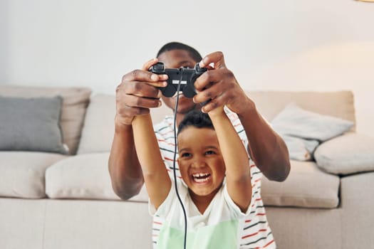 With joysticks in hands. African american father with his young son at home.