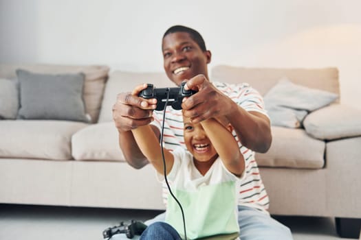 With joysticks in hands. African american father with his young son at home.