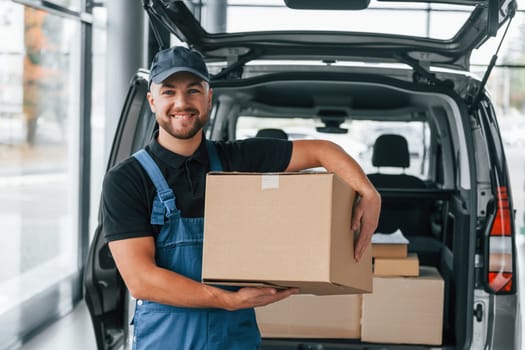 Holding box. Delivery man in uniform is indoors with car and with order.