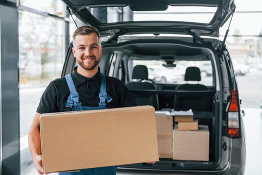 Holding box. Delivery man in uniform is indoors with car and with order.