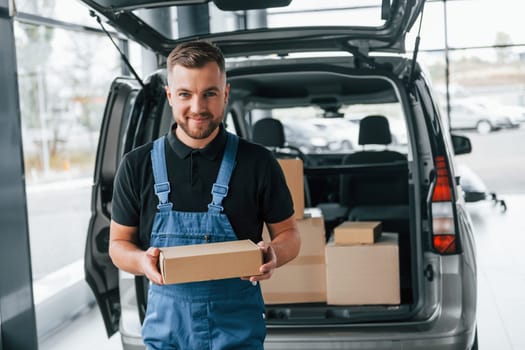 Holding box. Delivery man in uniform is indoors with car and with order.