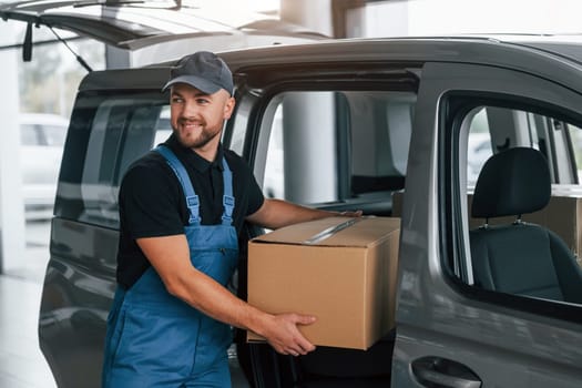 With box in hands. Delivery man in uniform is indoors with car and with order.