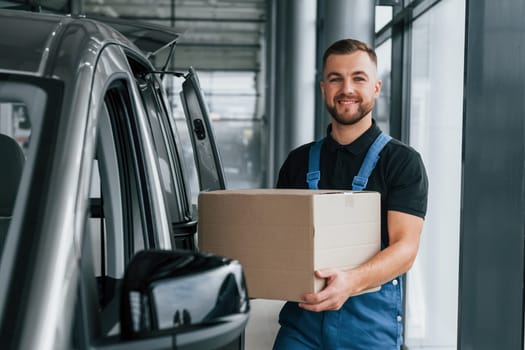 With box in hands. Delivery man in uniform is indoors with car and with order.
