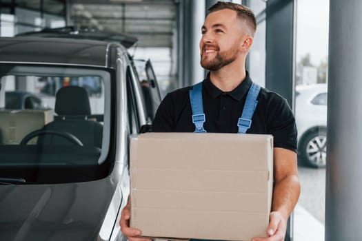 With box in hands. Delivery man in uniform is indoors with car and with order.