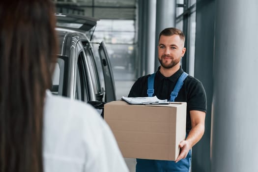 Woman waits for her order. Delivery man in uniform is indoors with car.