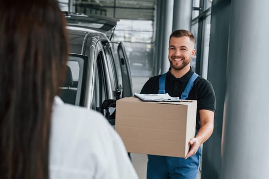 Woman waits for her order. Delivery man in uniform is indoors with car.