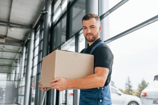 Front view. Delivery man in uniform is indoors with car and with order.