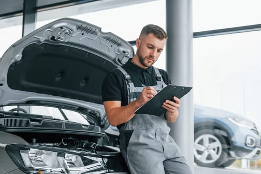 With notepad. Man in uniform is working in the autosalon at daytime.