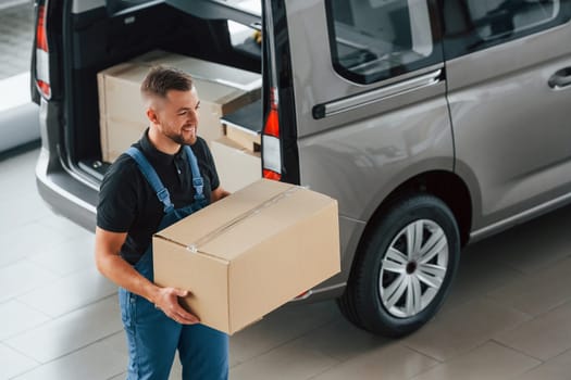 Walking and holding box in hands. Delivery man in uniform is indoors with car and with order.