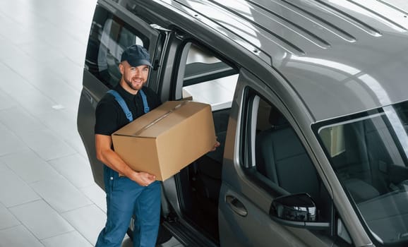 Walking and holding box in hands. Delivery man in uniform is indoors with car and with order.