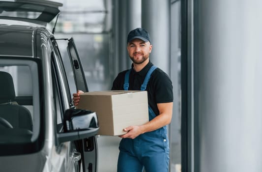 In blue colored clothes. Delivery man in uniform is indoors with car and with order.